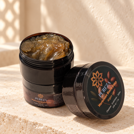 An open black jar of Pureza Moroccan Black Soap showing the brownish-gold gel inside, next to its lid which displays the brand logo and product name, set on a textured beige stone surface.