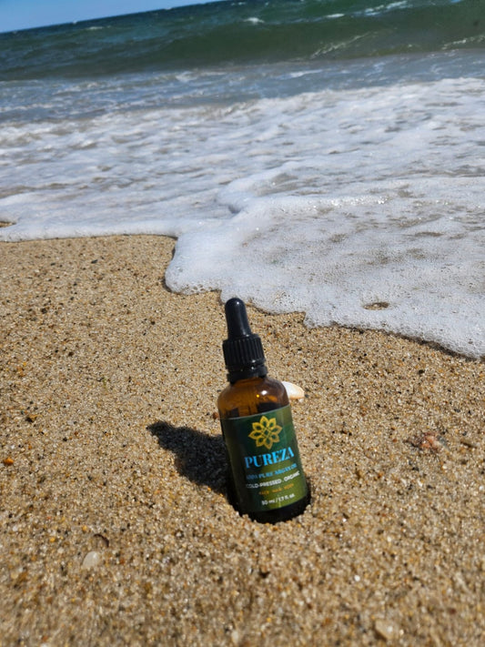 An Argan oil PUREZA bottle stands on sandy beach shore with ocean waves in the background.