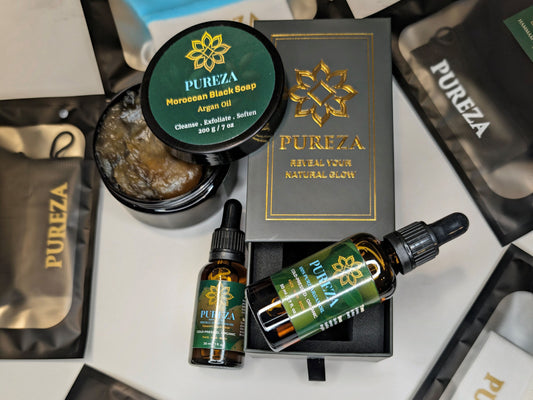 A collection of PUREZA natural skincare products arranged on a white surface, including a Moroccan black soap jar, an argan oil dropper bottle, a prickly pear seed oil bottle, and branded kessa gloves